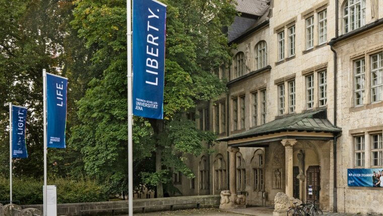 Blick auf das Universitätshauptgebäude mit Fahnen Light, Life, Liberty